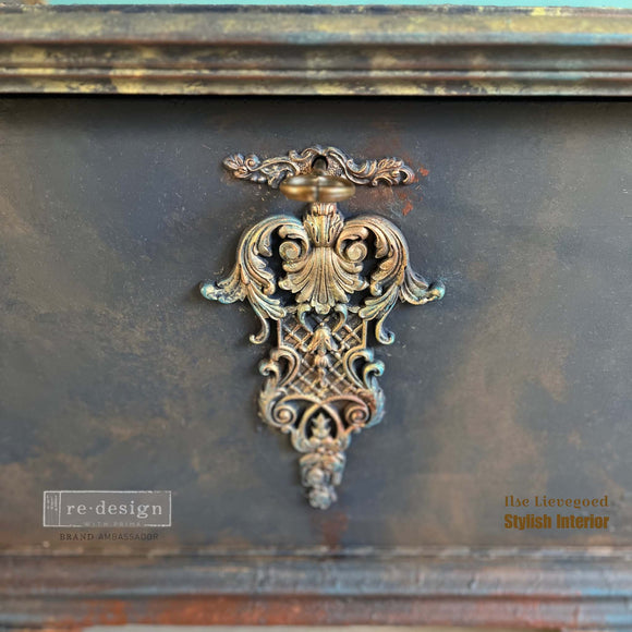 A close-up of ReDesign with Prima's Regal Finasse Decor Poly Casting painted in gold by Ilse Lievegoed is shown against a mottled gray piece of furniture.