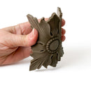 A hand is shown flexing a bendable furniture applique featuring a design of a scalloped edge square beneath a 4 petaled flower shaped flourish against a white background.