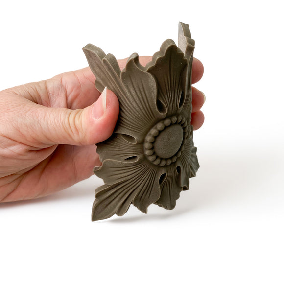 A hand is shown flexing a bendable furniture applique featuring a design of a scalloped edge square beneath a 4 petaled flower shaped flourish against a white background.