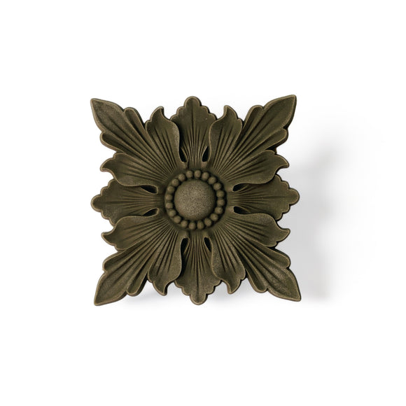 A bendable furniture applique featuring a design of a scalloped edge square beneath a 4 petaled flower shaped flourish is against a white background.