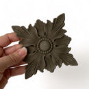 A hand is shown holding a bendable furniture applique featuring a design of a scalloped edge square beneath a 4 petaled flower shaped flourish against a white background.