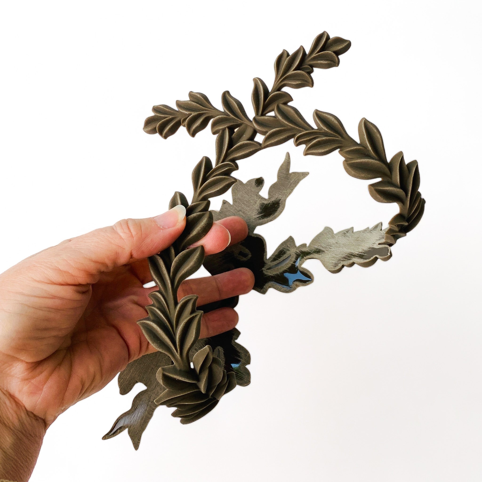 A hand is shown holding a curved bendable furniture applique of leafy sprigs intertwining in an oval frame with the appearance of a ribbon tie against a white background.