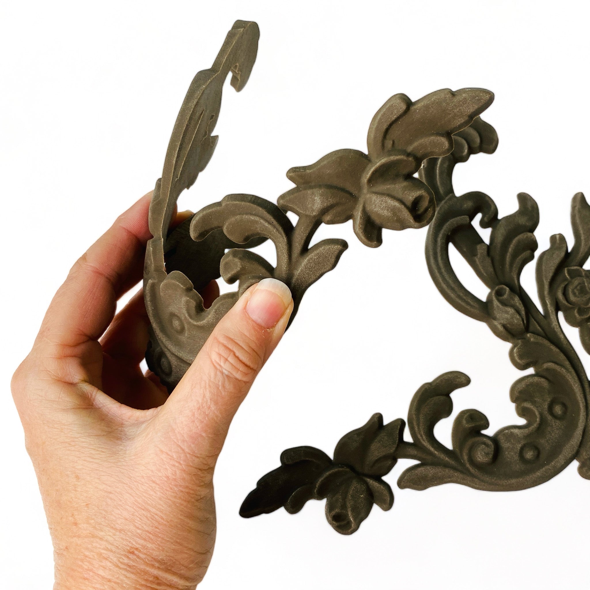 A hand is shown holding and flexing 1 of 2 bendable furniture appliques that feature corner flourishes, adorned with delicate leaves and flowers against a white background.