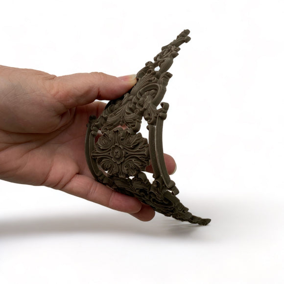 A hand is shown flexing a bendable furniture applique that features scrolling flourishes against a white background.