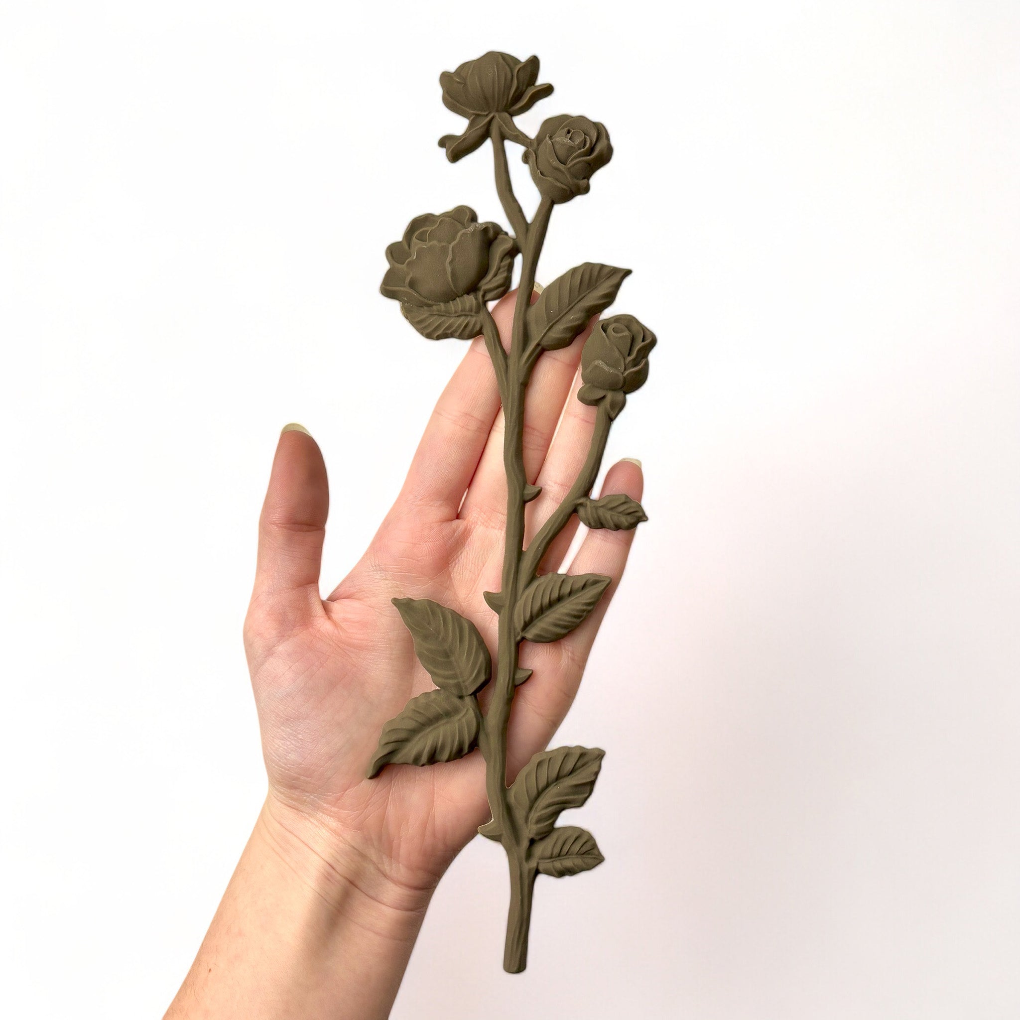 A hand is shown holding a brown colored decor poly bendable furniture applique of long stem roses against a white background.