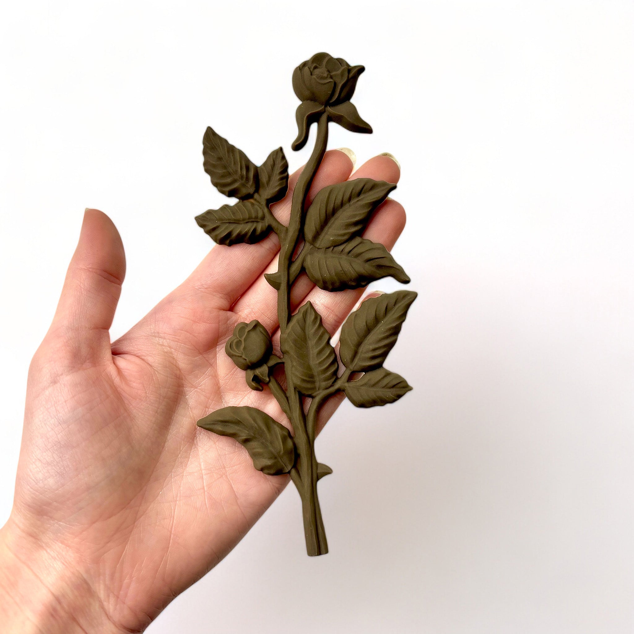 A hand is shown holding a brown colored decor poly bendable furniture applique of long stem roses against a white background.