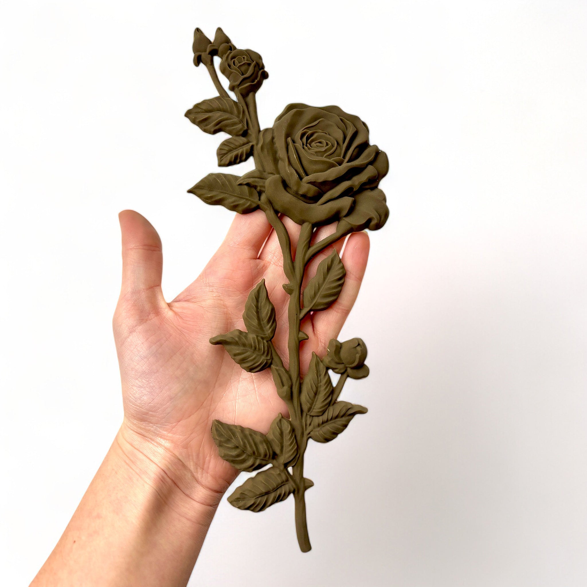 A hand is shown holding a brown colored decor poly bendable furniture applique of long stem roses against a white background.