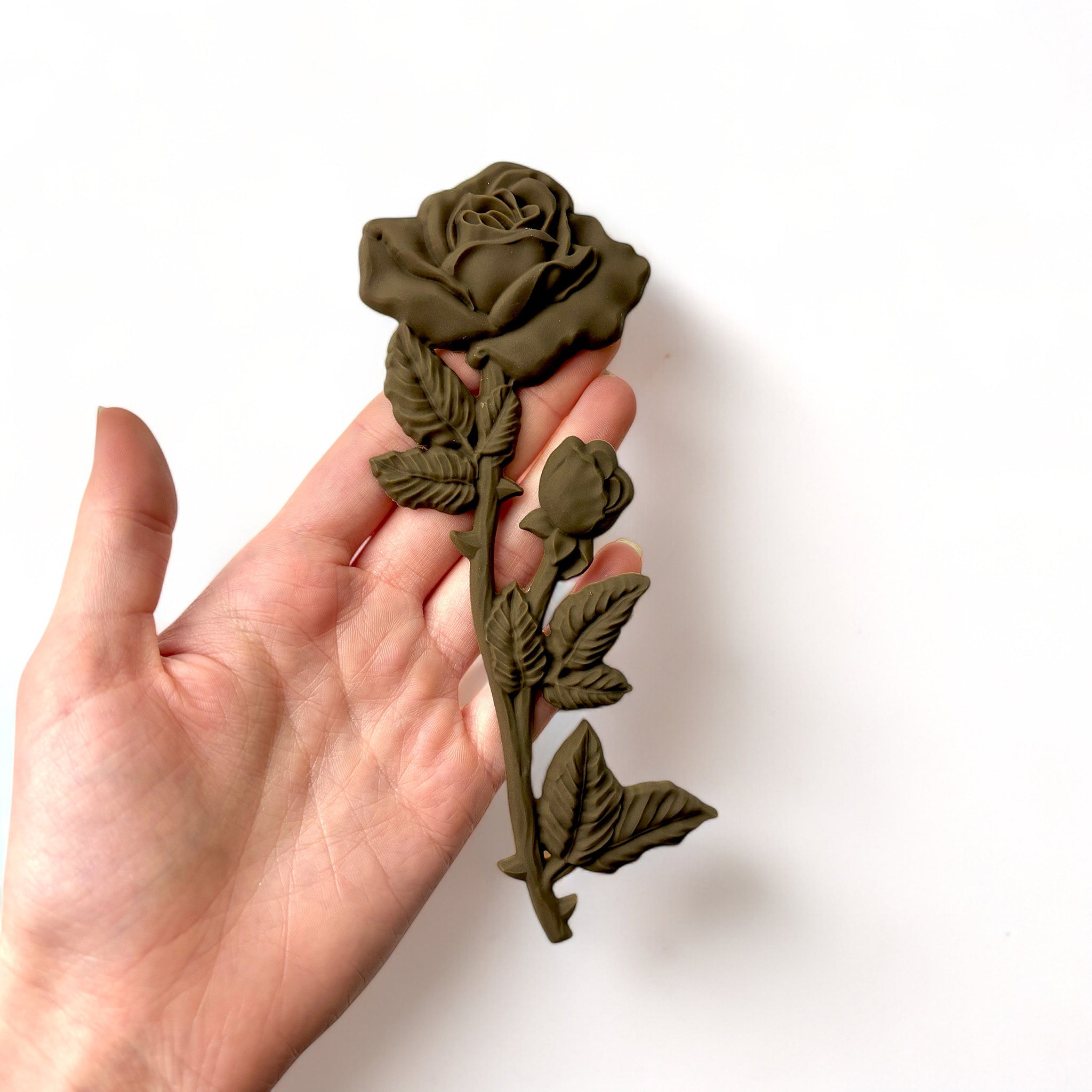 A hand is shown holding a brown colored decor poly bendable furniture applique of long stem roses against a white background.