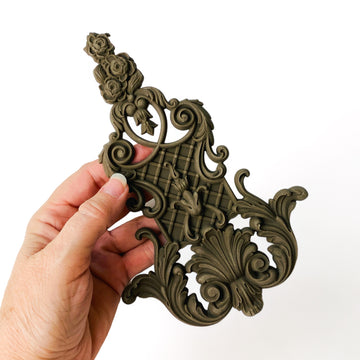 A hand is shown holding a bendable furniture applique that features intricate flourishes, a lattice center, and floral accents against a white background.