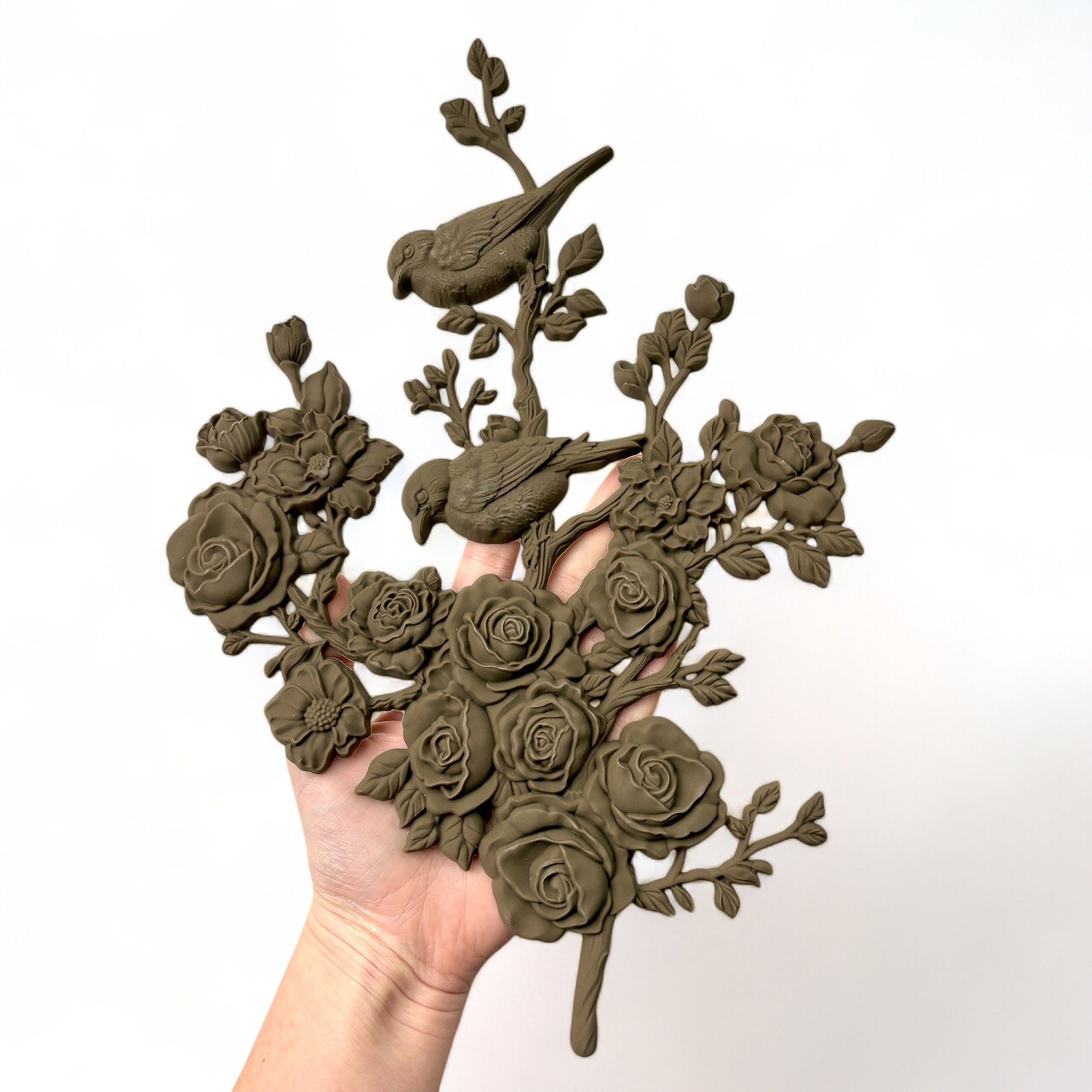 A hand is shown holding a brown colored bendable furniture applique of 2 birds perched on a branch of blooming roses and flowers against a white background.