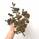 A hand is shown holding a brown colored bendable furniture applique of 2 birds perched on a branch of blooming roses and flowers against a white background. 