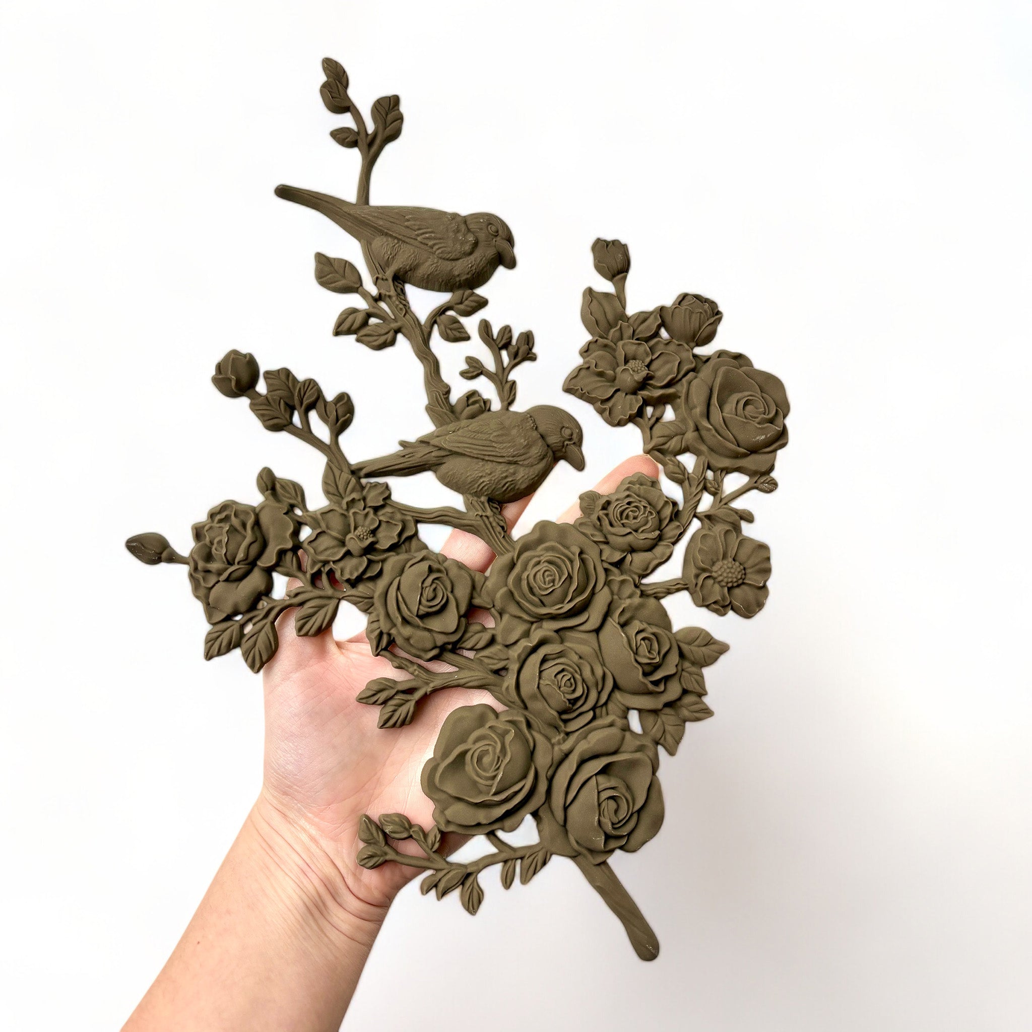 A hand is shown holding a brown colored bendable furniture applique of 2 birds perched on a branch of blooming roses and flowers against a white background. 