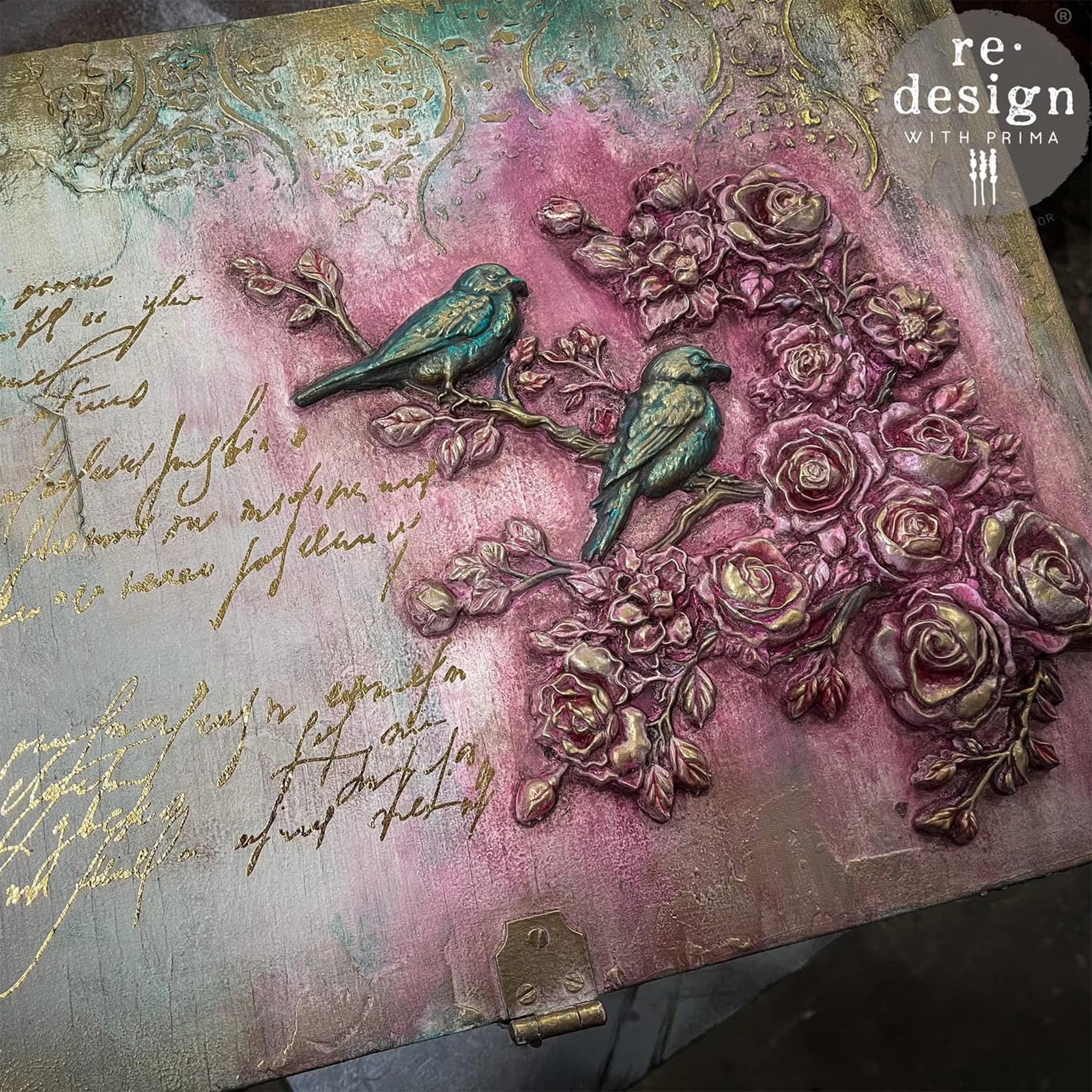 A close-up of a box painted with patina green and gold with gold foil script writing features ReDesign with Prima's Serene Perch Right Decor Poly painted in purple and gold on the lid.