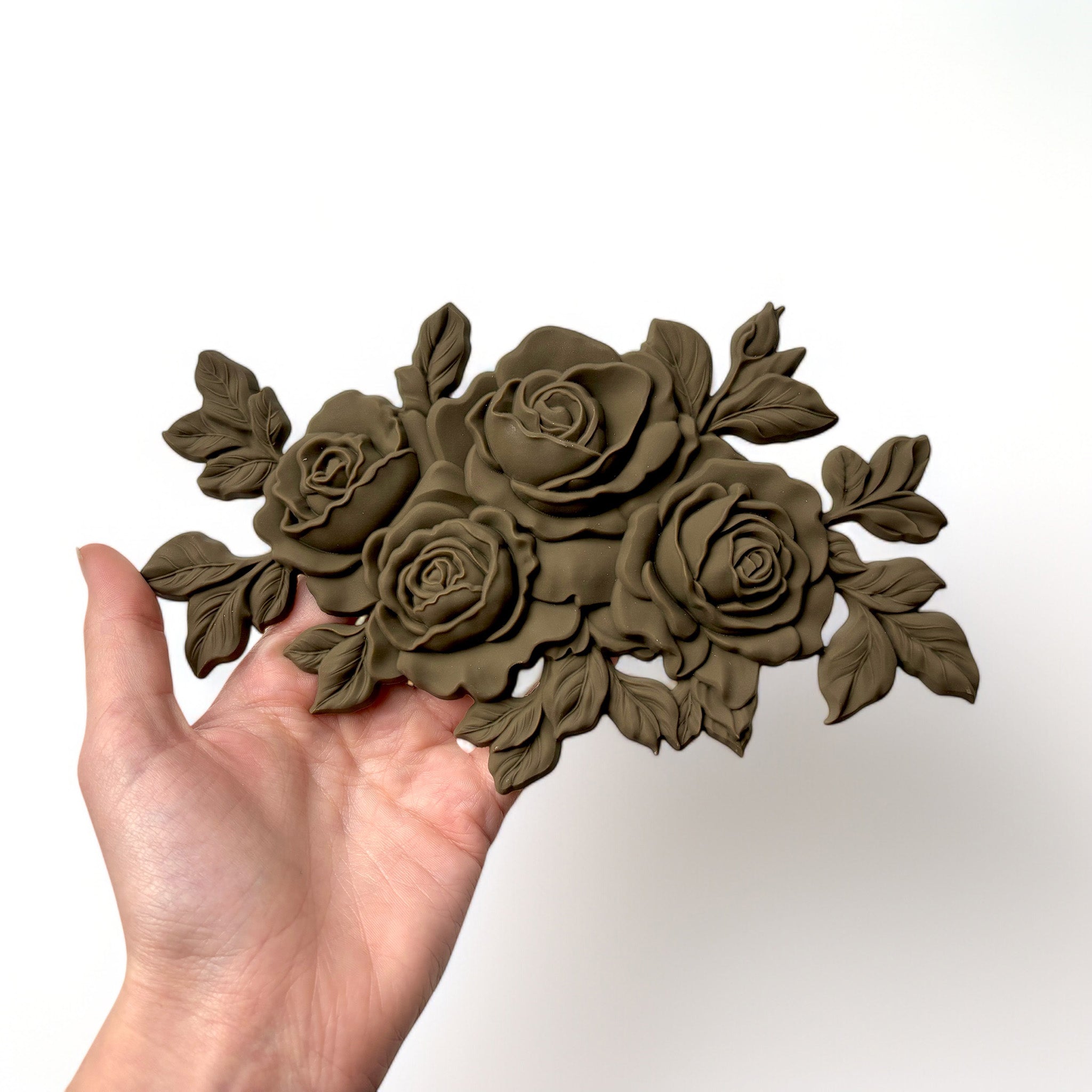 A hand is shown holding a brown colored bendable furniture applique of a small bouquet of 4 roses against a white background.
