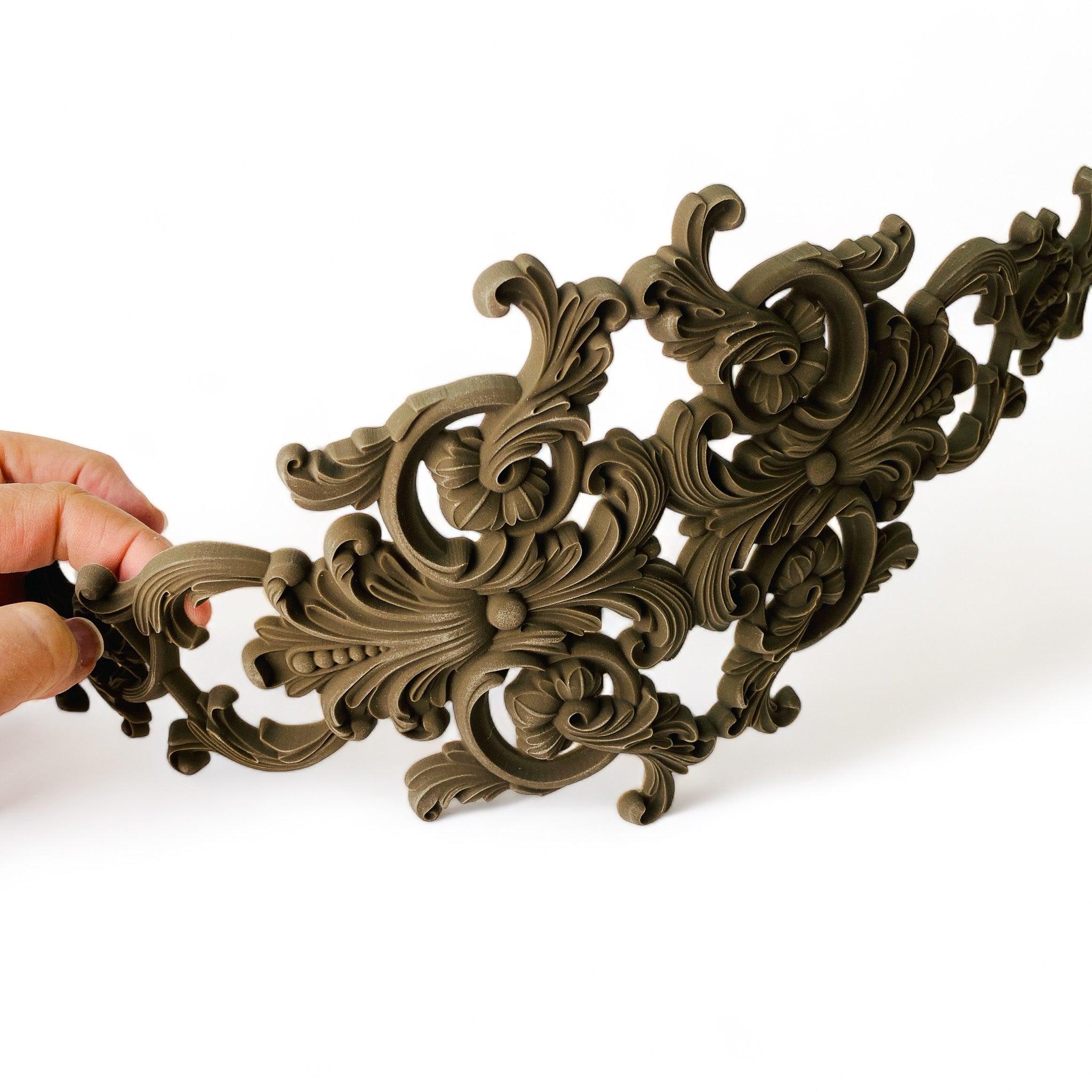 A hand holding a curved bendable furniture applique featuring scrolling ornate details is against a white background.
