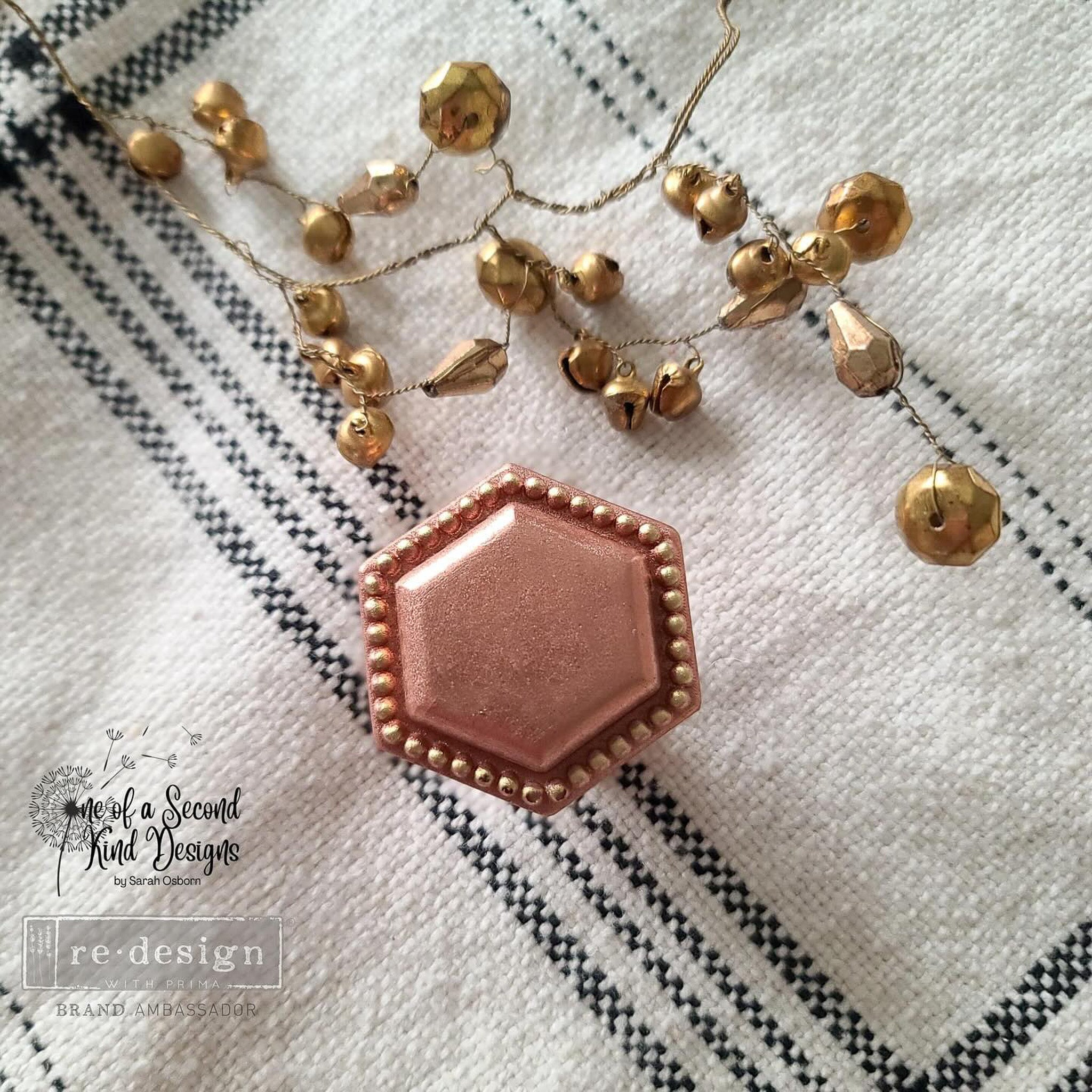 A rose gold colored, pearl-edged, hexagon-shaped silicone mold casting using ReDesign with Prima's Imperial Pearl 3D Knob mold created by One of a Second Kind Designs is sitting on a white napkin with black plaid. Small gold jingle bells on wire are above the knob.