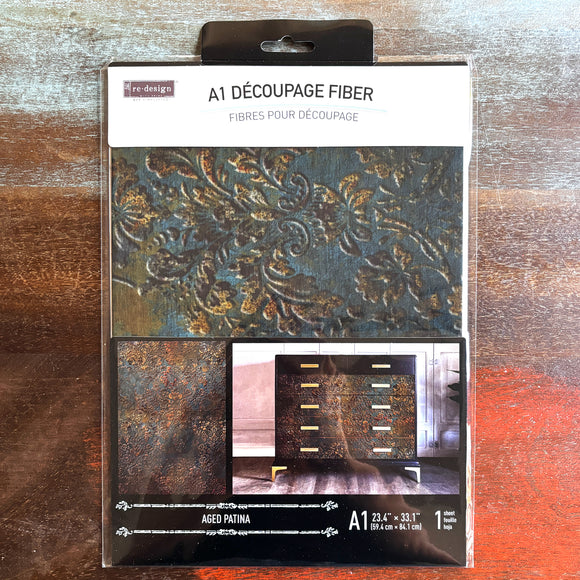 A package of ReDesign with Prima's Aged Patina A1 fiber paper is against a dark wood background.