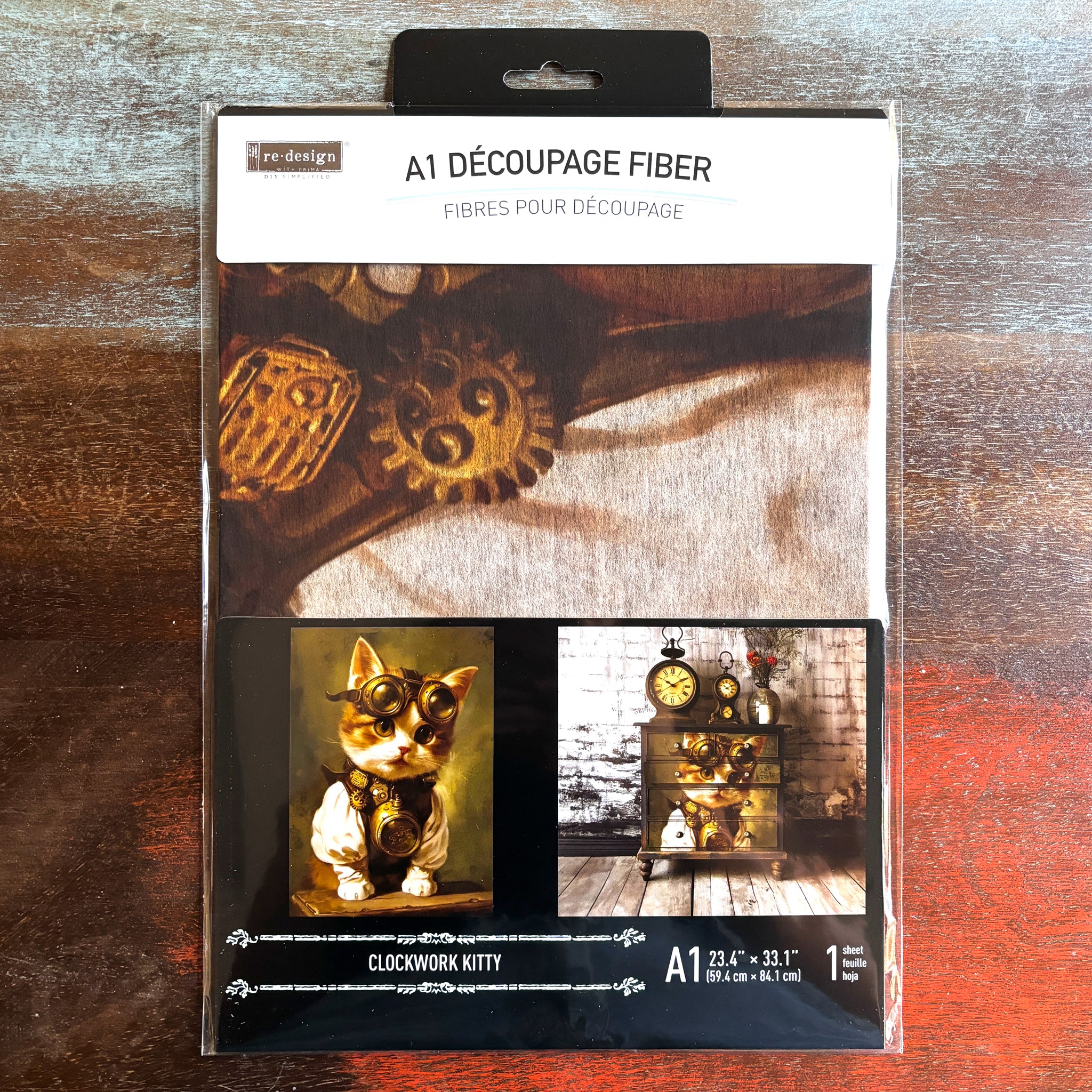 A package of ReDesign with Prima's Clockwork Kitty A1 fiber paper is against a dark wood background.