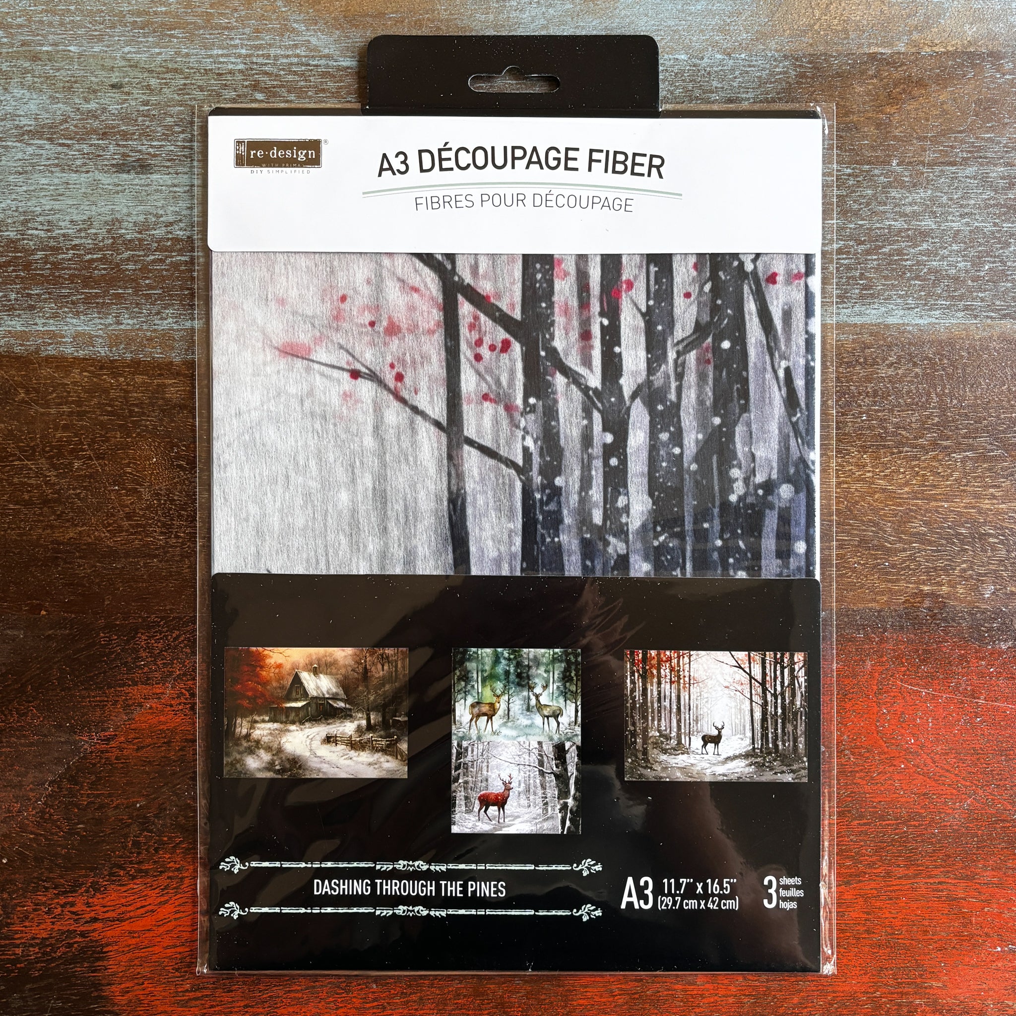 A package of ReDesign with Prima's Dashing Through the Pines A3 fiber paper 3 pack is against a dark wood background.