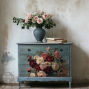 Decorative chest of drawers with floral design and a vase of flowers on top against a textured wall.
