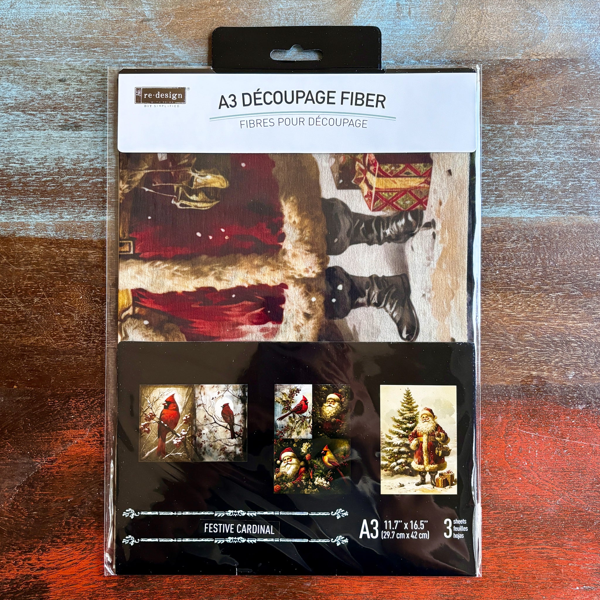 A package of ReDesign with Prima's Festive Cardinal A3 fiber paper 3 pack is against a dark wood background.