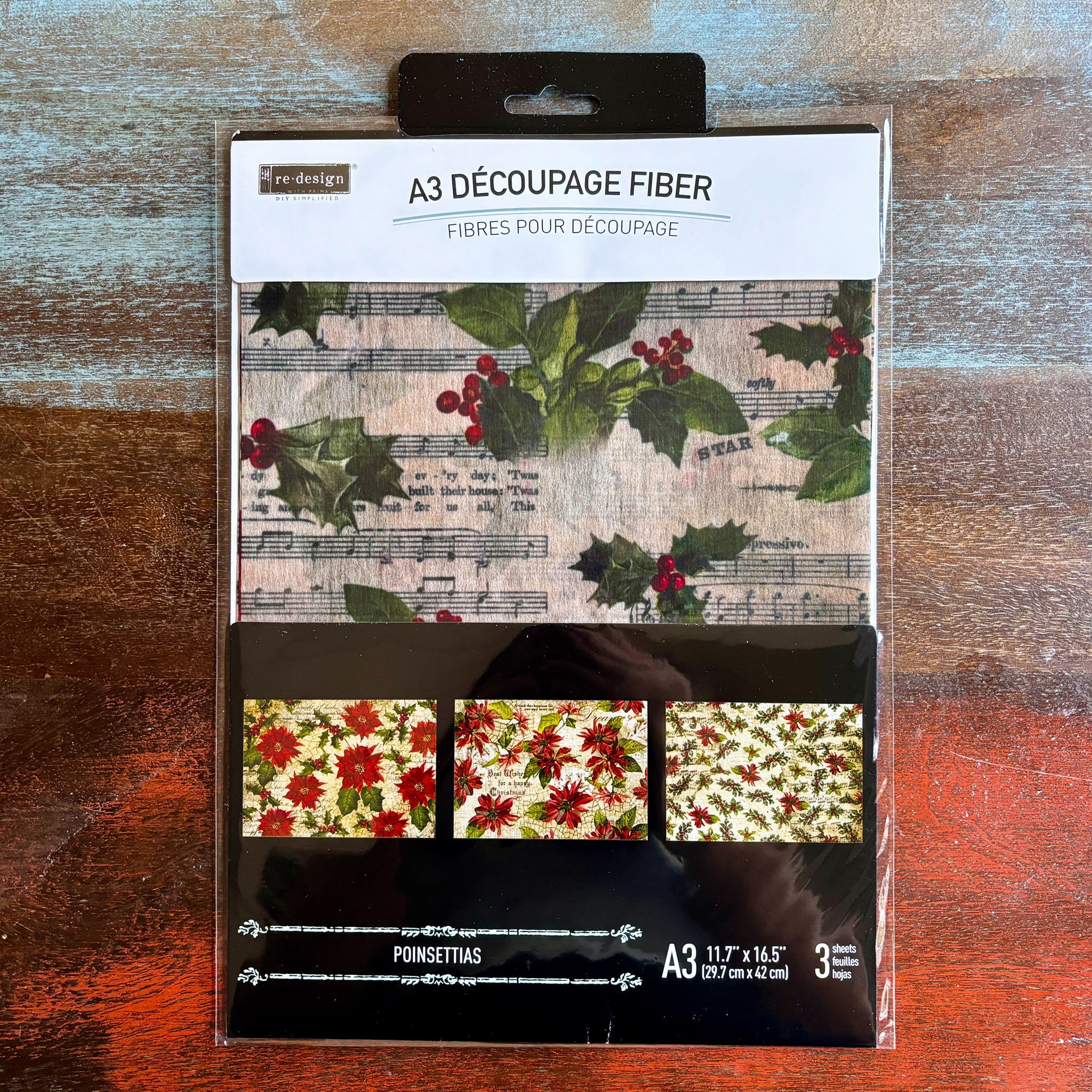 A package of ReDesign with Prima's Poinsettias A3 fiber paper 3 pack is against a dark wood background.