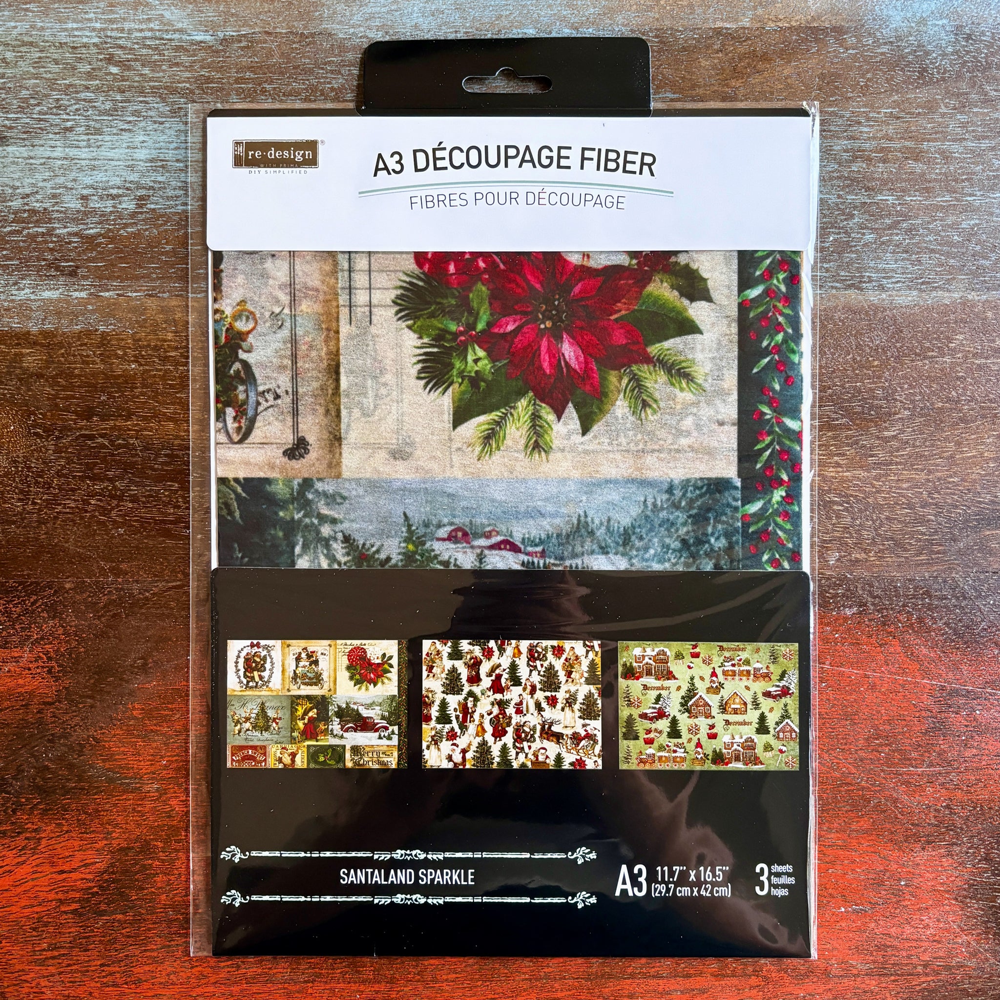 A package of ReDesign with Prima's Santaland Sparkle A3 fiber paper 3 pack is against a dark wood background.