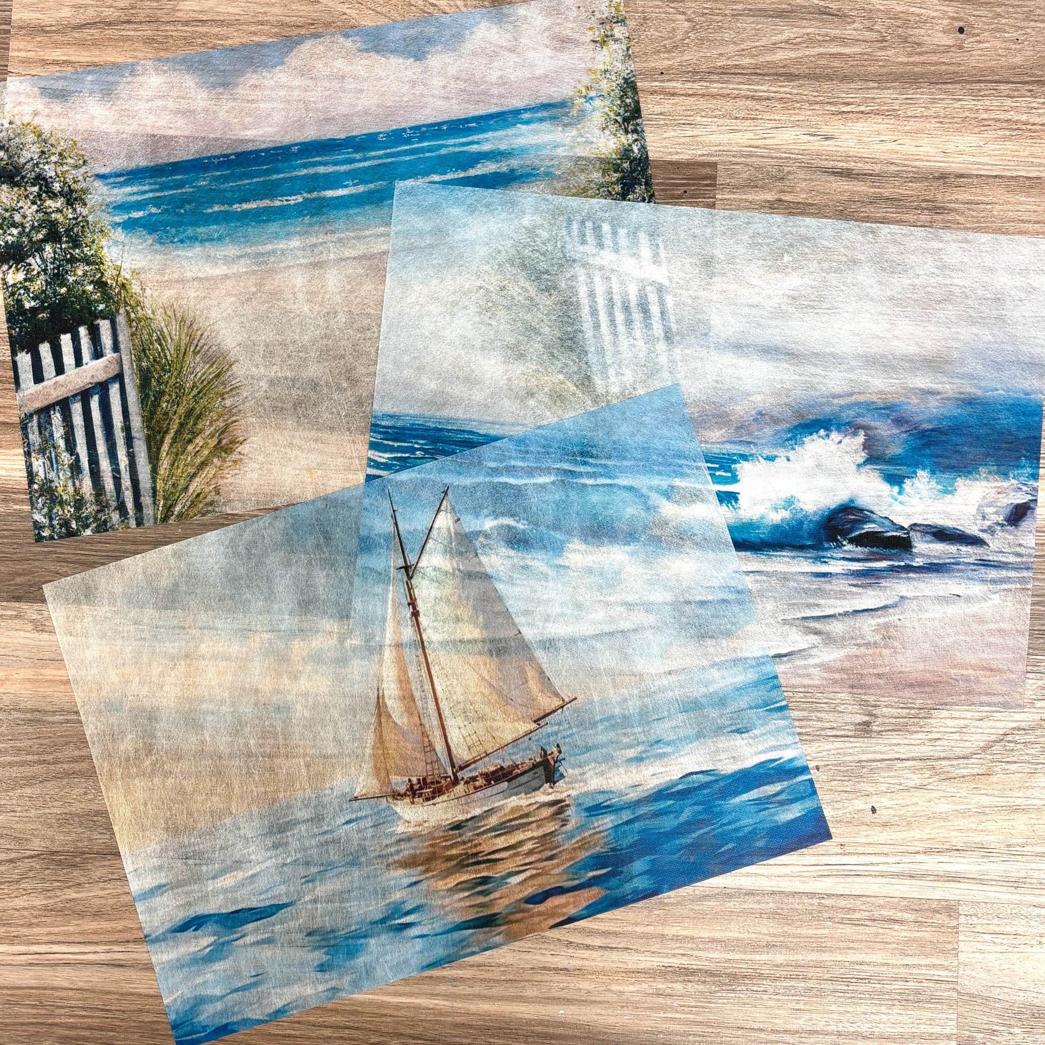 Three sheets of A3 fiber papers featuring scenes of a quiet beach, crashing waves, and a sailboat are against a light wood background.