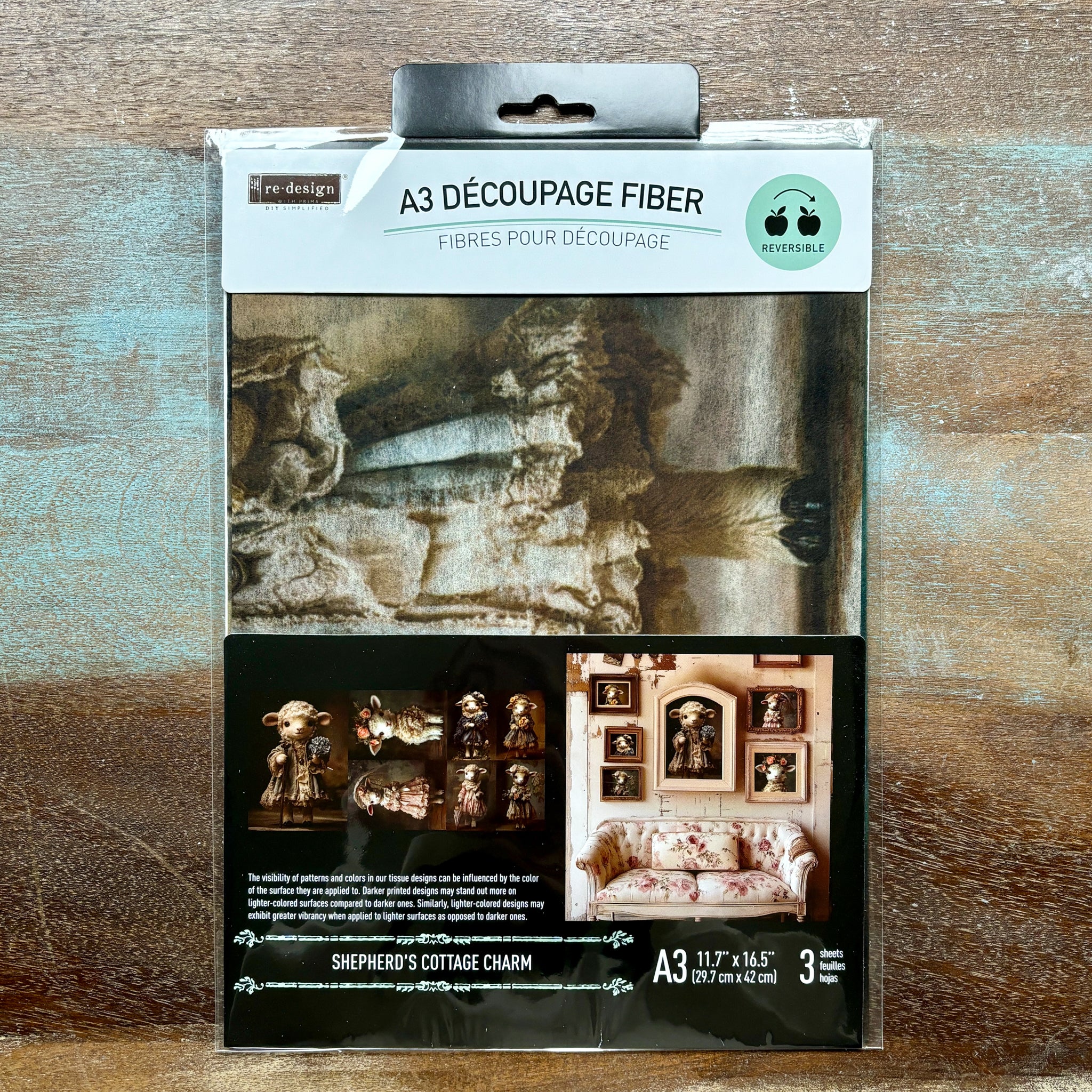 A package of ReDesign with Prima's Shepherd's Cottage Charm A3 fiber paper 3 pack is against a wood background.