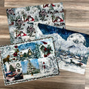 Three sheets of A3 fiber paper featuring winter scenes of villages, snow covered mountains, and collages of Christmas trees, snowmen, and red cardinal birds on a birdhouse are against a light wood background.