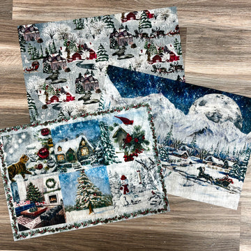 Three sheets of A3 fiber paper featuring winter scenes of villages, snow covered mountains, and collages of Christmas trees, snowmen, and red cardinal birds on a birdhouse are against a light wood background.