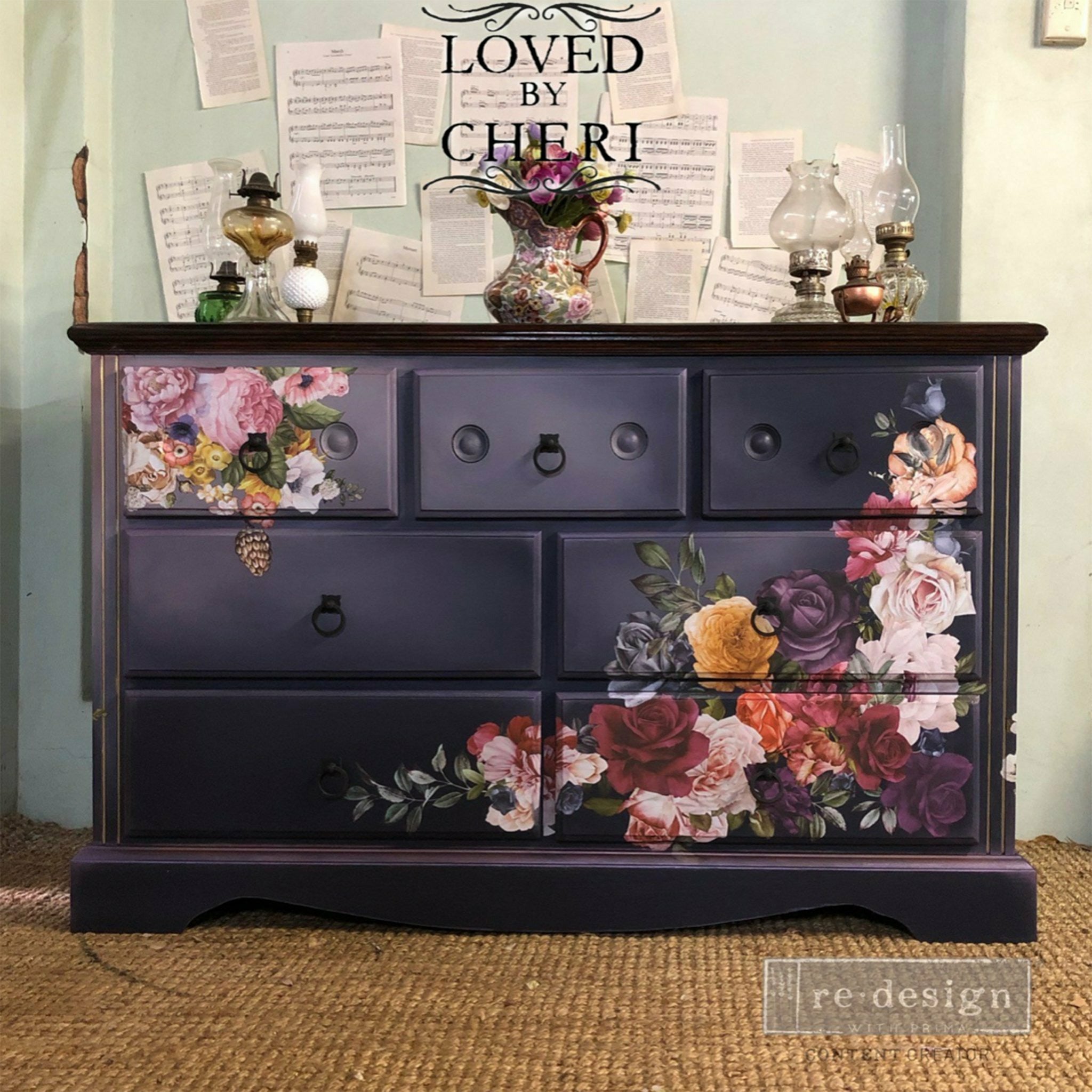 A large 7-drawer dresser refurbished by Loved by Cheri is painted a blend of midnight blue and gray and features ReDesign with Prima's Beautiful Things rub on transfer on the front top left and bottom right corners.