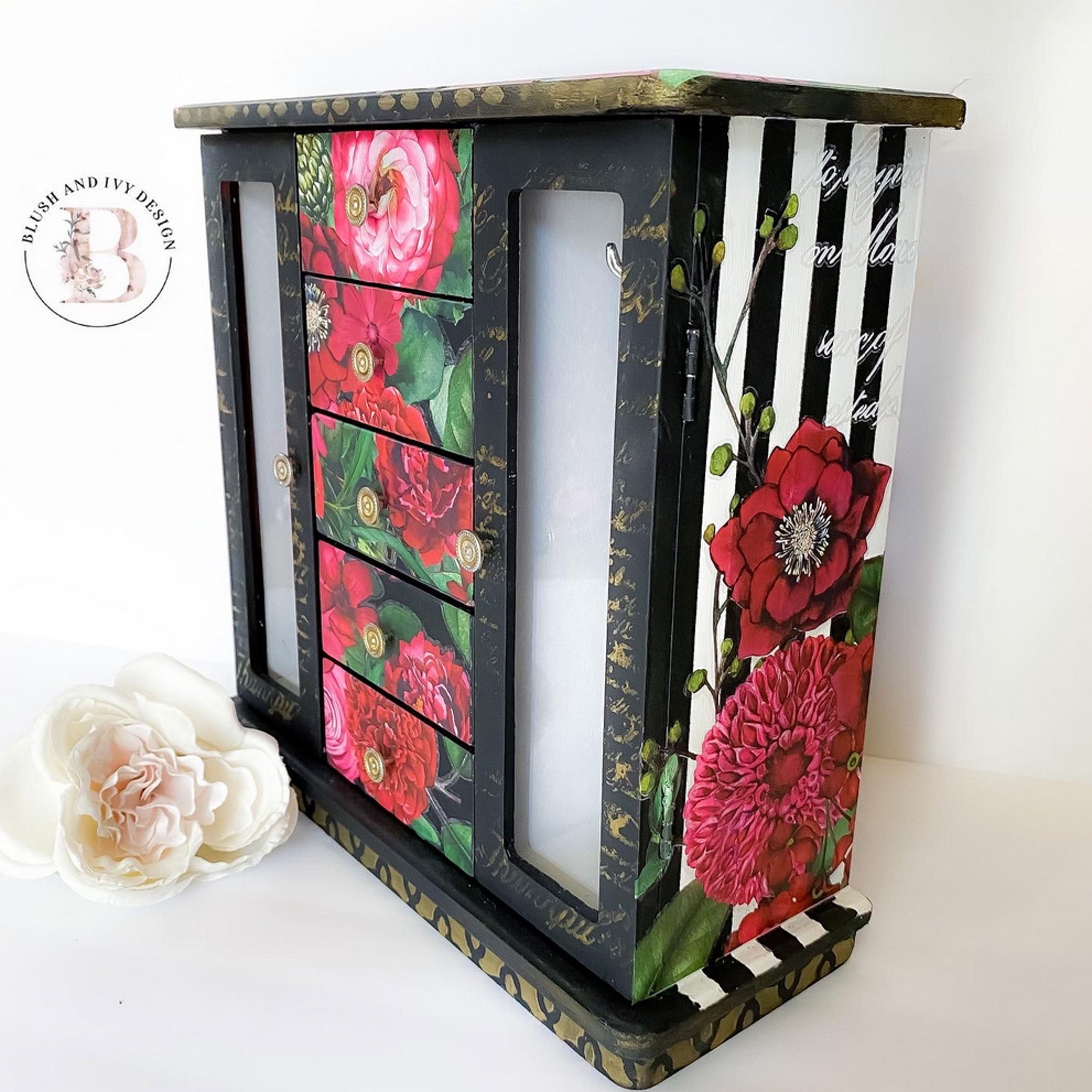 A corner side view of a table top jewelry armoire refurbished by Blush and Ivy Design is painted black with white stripes on the sides and features ReDesign with Prima's Earthly Delights rub on transfer on it.