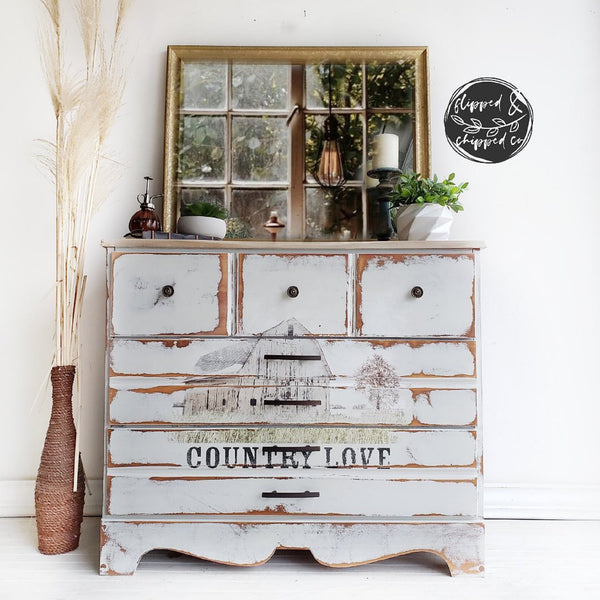 A vintage dresser refurbished by Flipped & Chipped Co. is painted distressed white and features ReDesign with Prima's Out on the Farm transfer on its drawers.