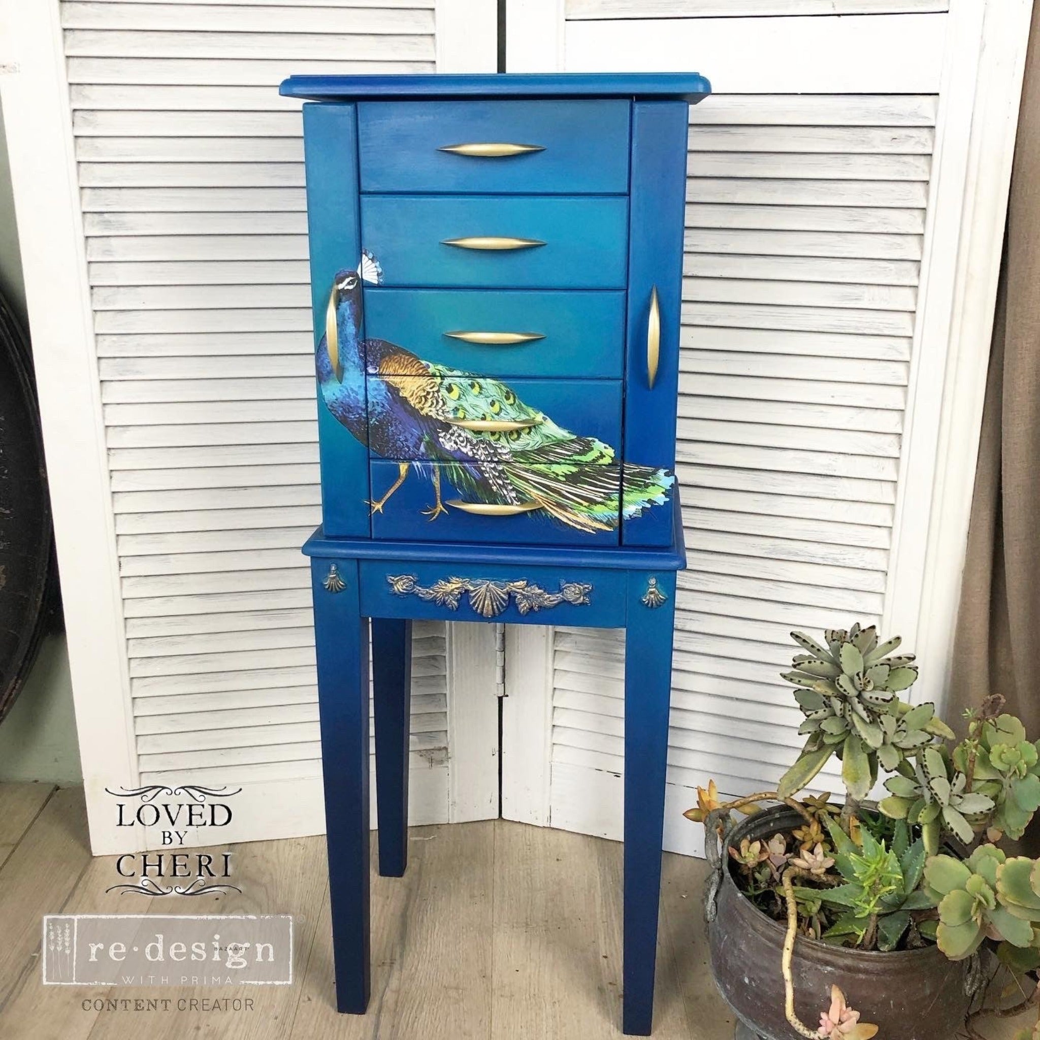 A vintage 5-drawer standing jewelry armoire refurbished by Loved by Cheri is painted blue and features ReDesign with Prima's Peacock Dreams transfer on the front.