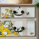 Close-up of a painted dresser showing Sunlit Serenade by ReDesign with Prima—yellow florals, green vines, and bee details applied across two drawers with dark vintage hardware