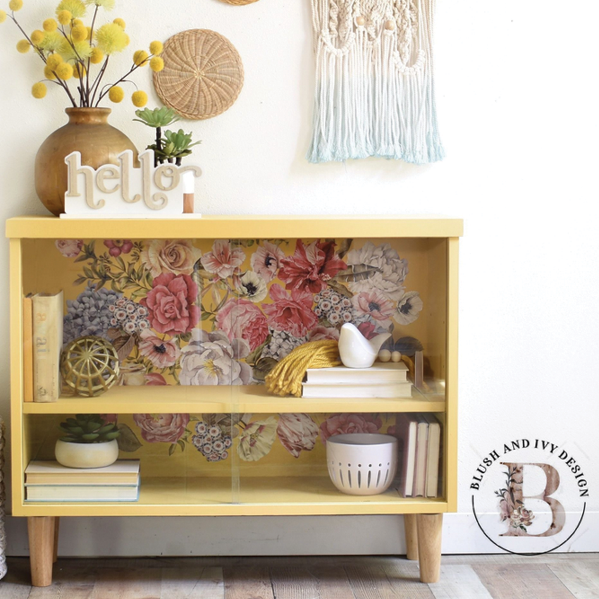 A small dresser turned into a bookcase refurbished by Blush and Ivy Design is painted pale yellow and features ReDesign with Prima's Wondrous Floral 2 transfer against its backboard.