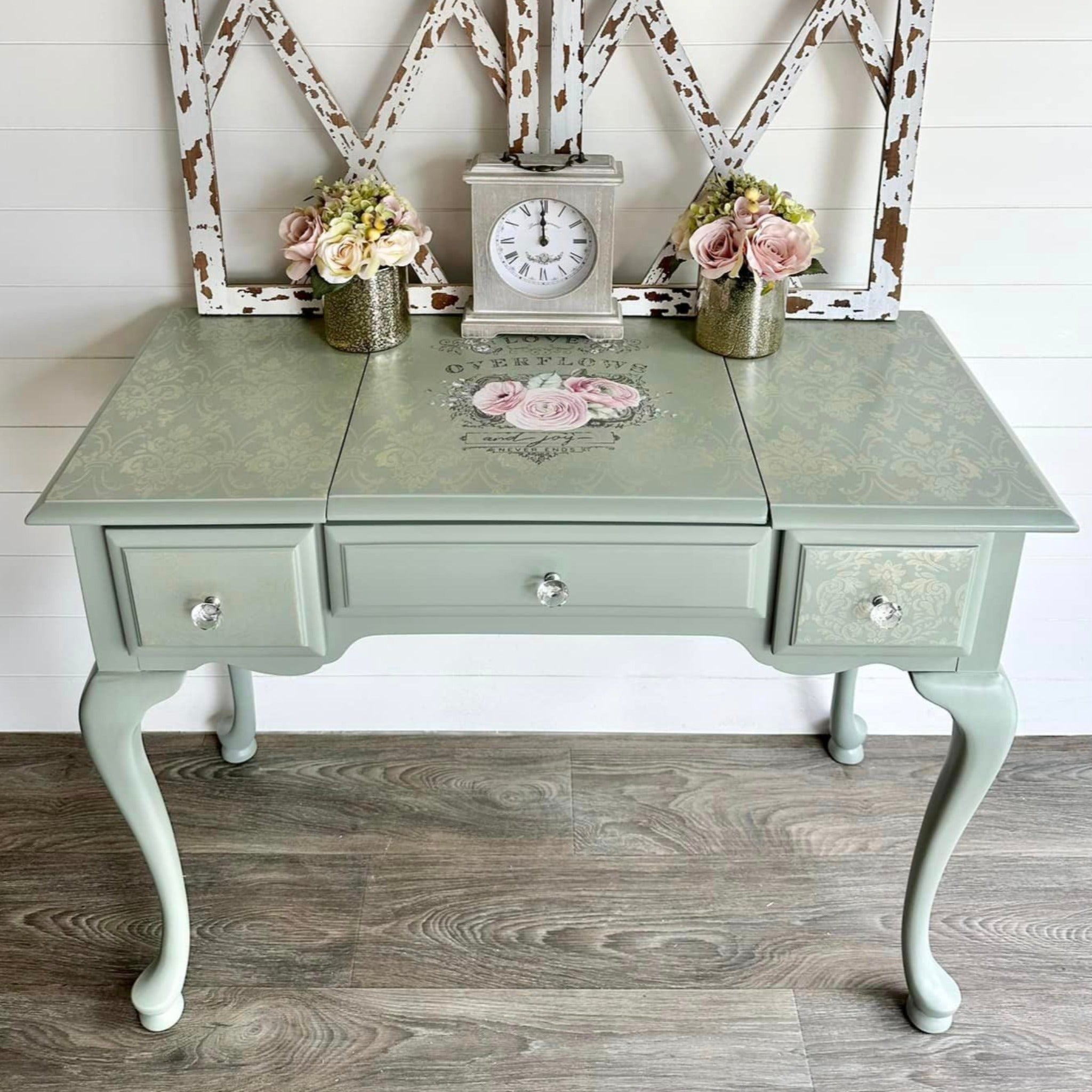 A vintage vanity desk is painted light sage green and features ReDesign with Prima's Overflowing love transfer on its top lift-up lid.