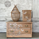 Wooden dresser with decorative drawers and a birdcage against a textured wall.