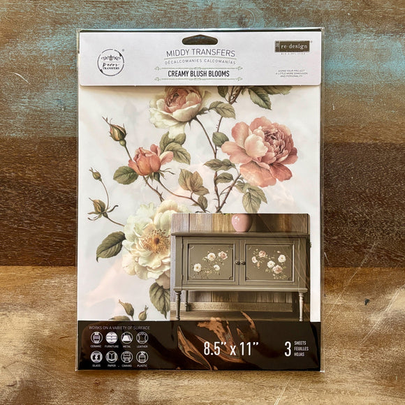 A package of ReDesign with Prima's Creamy Blush Blooms small rub-on transfers is against a wood background.