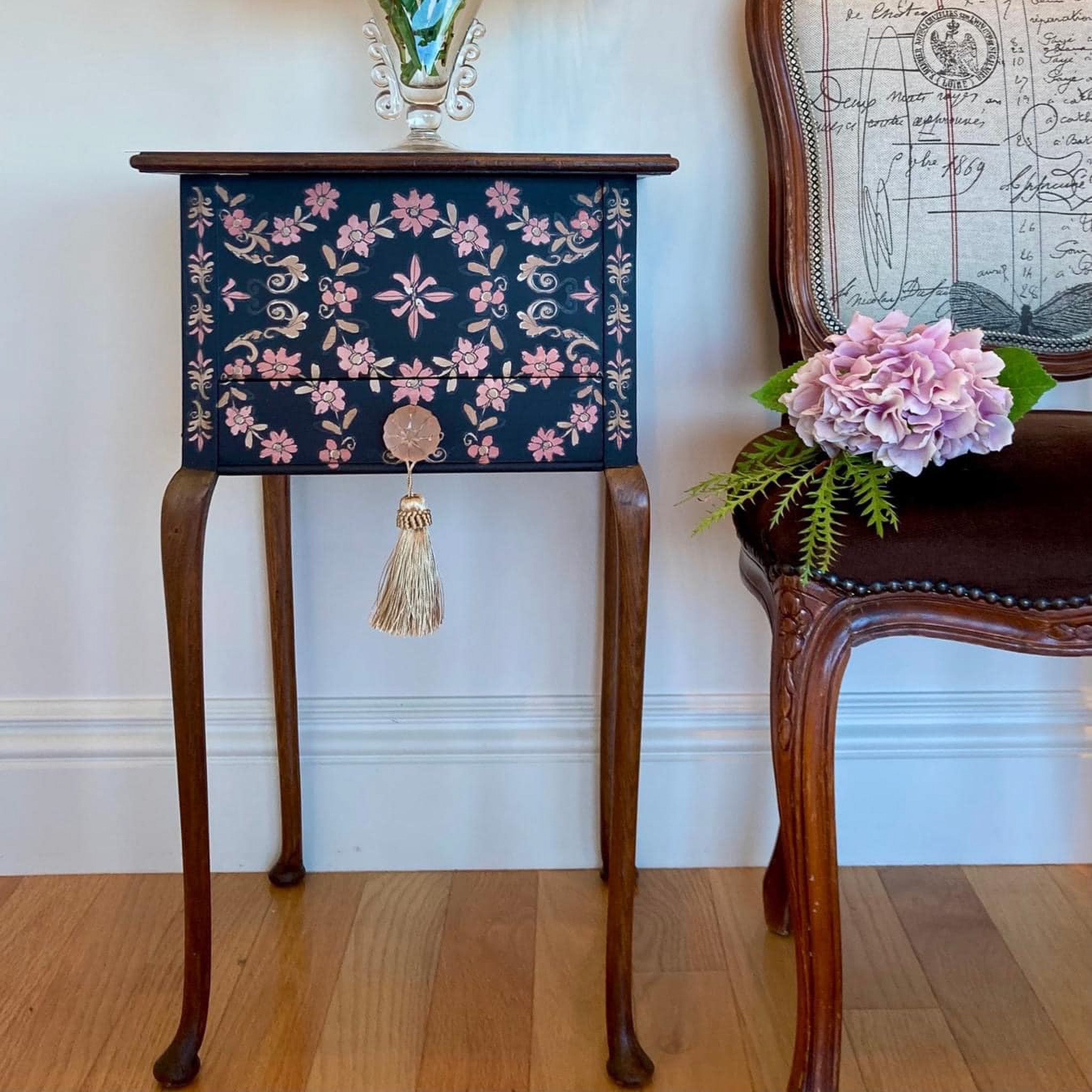A small vintage side table is painted dark blue with natural wood accents and features ReDesign with Prima's Annie Sloan Flower Garland rub-on transfer on its drawer.