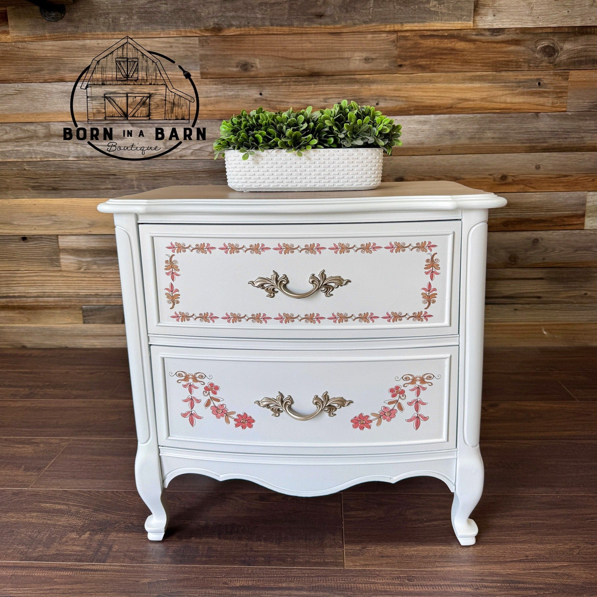 A small white 2-drawer nightstand refurbished by Born in a Barn Boutique features ReDesign with Prima's Annie Sloan Flower Garland rub-on transfer on the drawers.