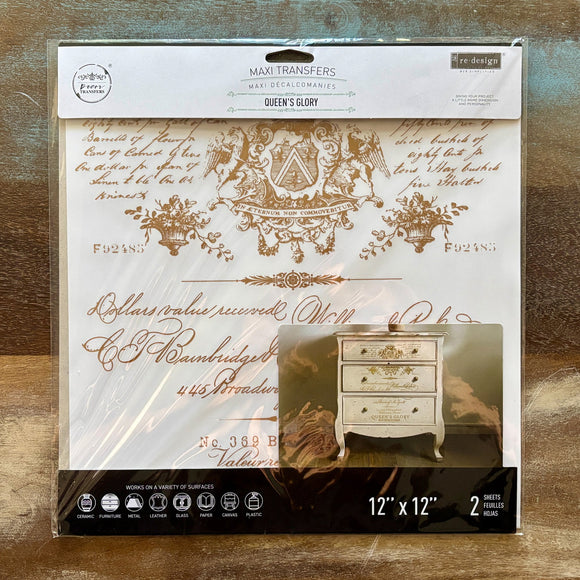 A package of ReDesign with Prima's Queen's Glory 12"x12" rub-on transfers are against a wood background.