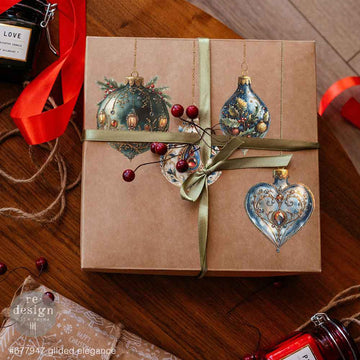 A brown square box with a gold ribbon features glass ornament designs from ReDesign with Prima's Gilded Elegance middy rub-on transfers on the lid.