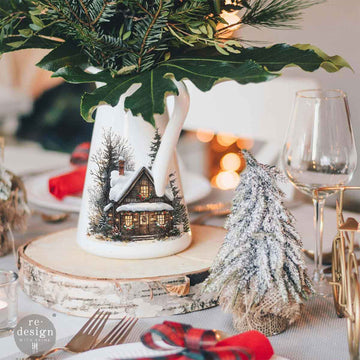 A white ceramic milk cream jug turned into a vase with winter greens in it features a design from ReDesign with Prima's Village of Wonder middy rub-on transfers.