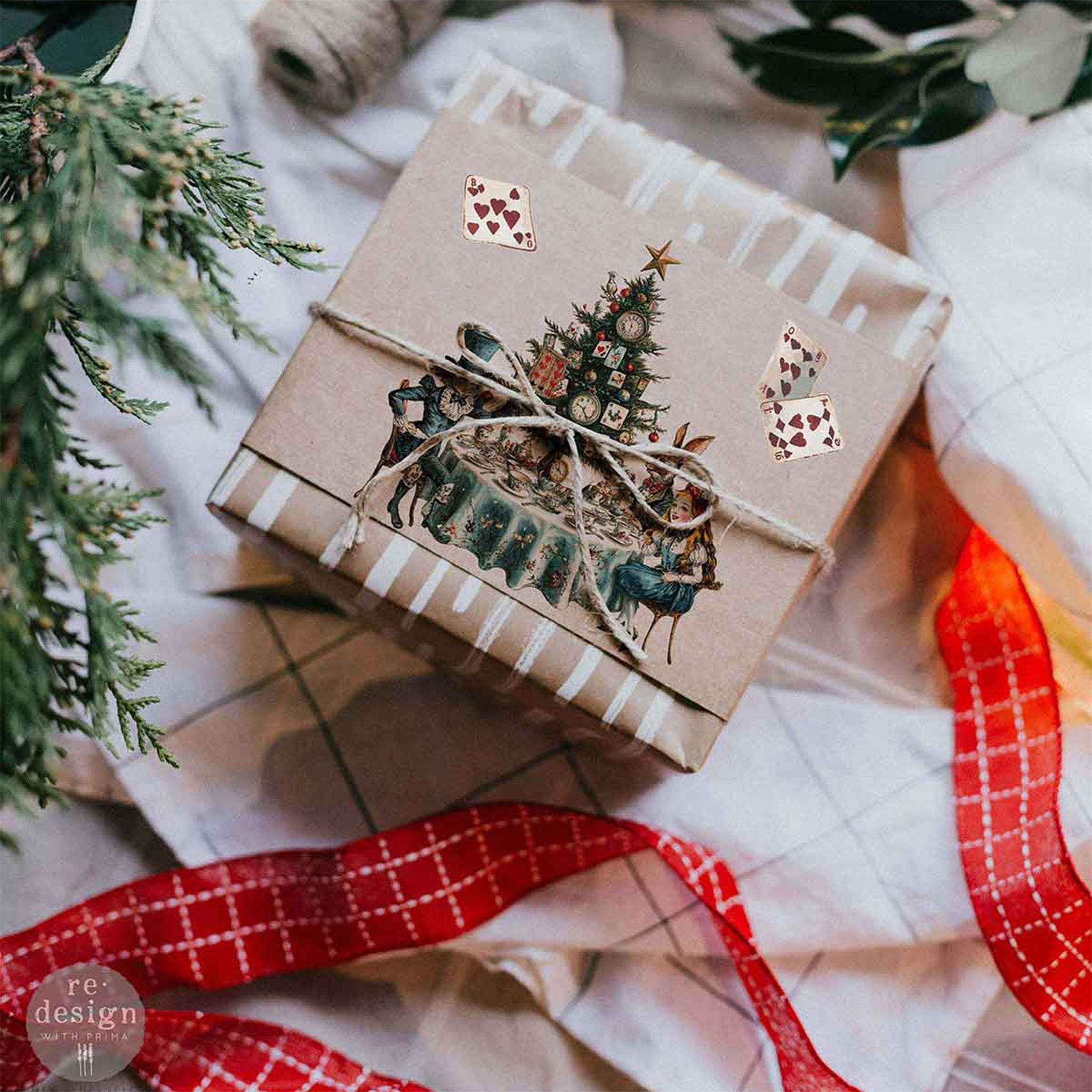 A brown paper wrapped gift nestled along red ribbon features a design from ReDesignwith Prima's Whimsical Holiday Dreams middy rub-on transfers.