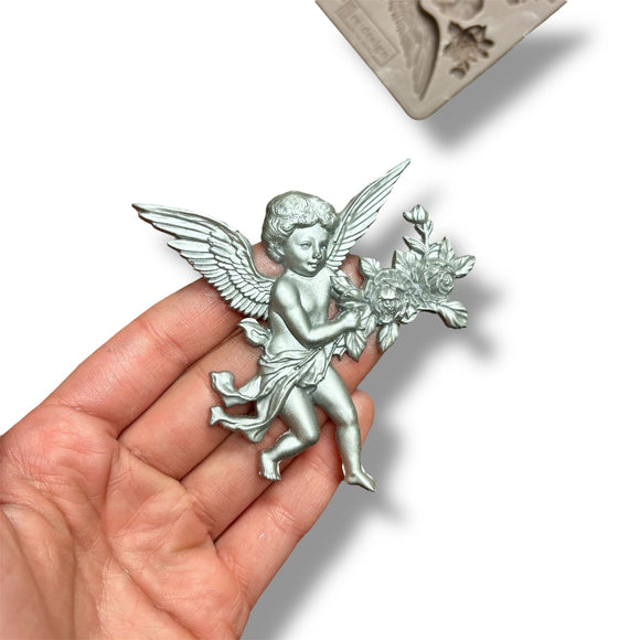 A hand against a white background is shown holding a silver colored casting of a cherub created from ReDesign with Prima's Angelic Aura silicone mold.