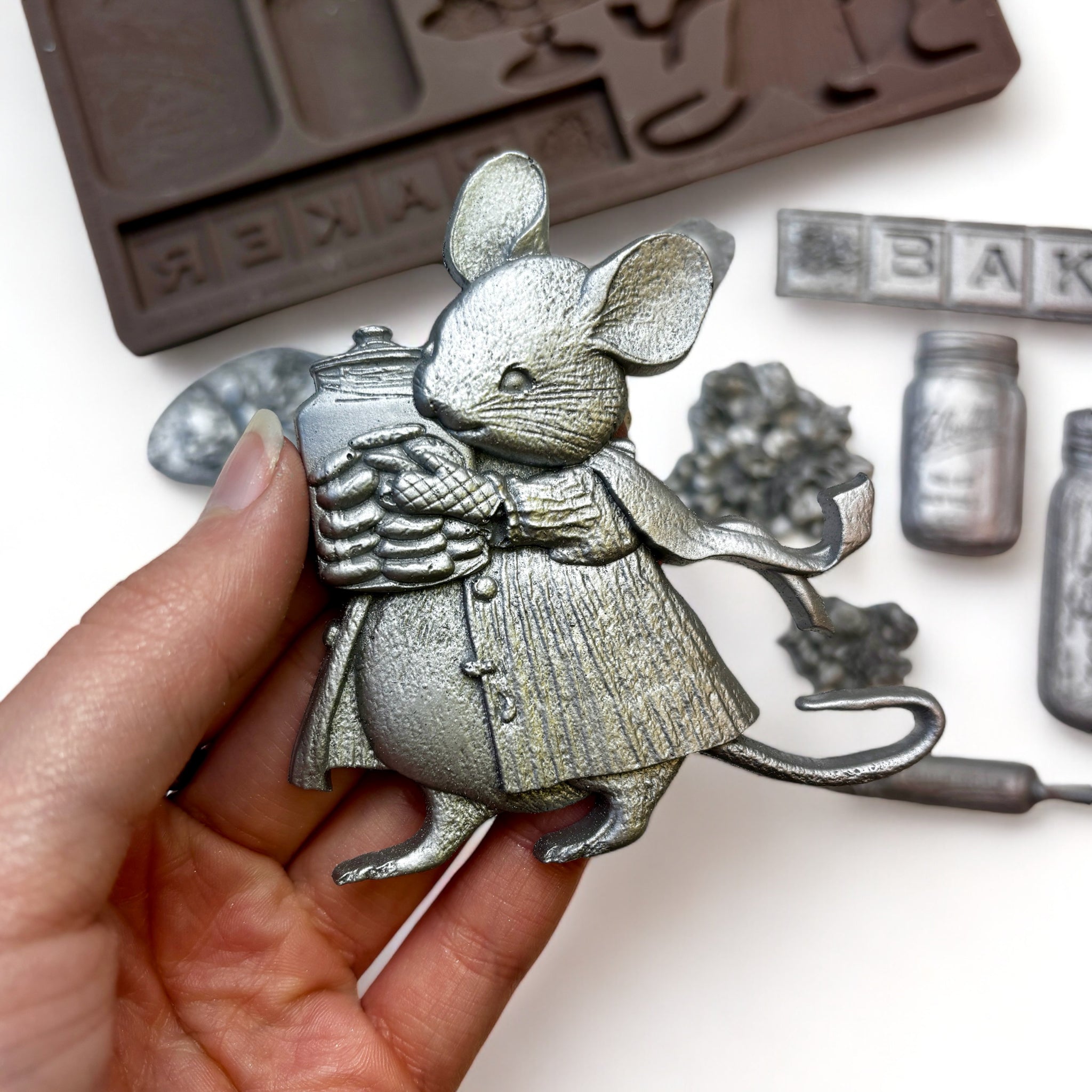 A brown silicone mold A silver colored castings of a small baker mouse with vintage kitchen baking items and 2 small floral bouquets are against a white background. A hand is shown holding the mouse casting.