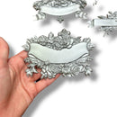 A hand is shown holding a silver colored casting of a banner surrounded by roses against a white background with 2 other castings created from ReDesign with Prima's Blooming Banners silicone mold.