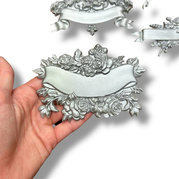 A hand is shown holding a silver colored casting of a banner surrounded by roses against a white background with 2 other castings created from ReDesign with Prima's Blooming Banners silicone mold.